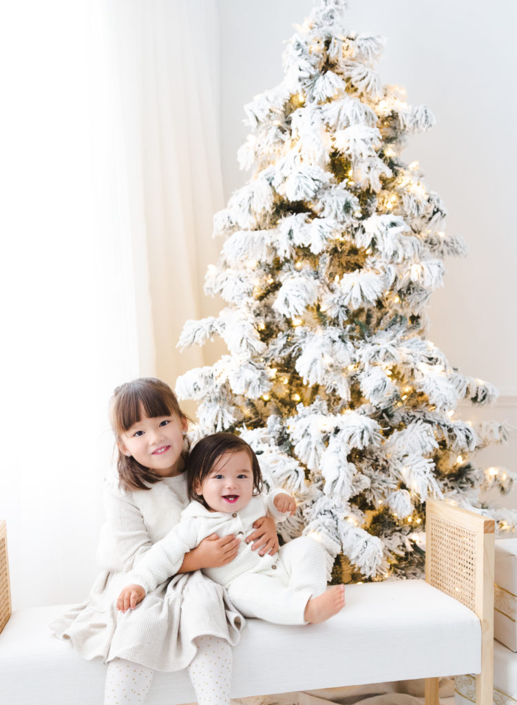 Two young children in white pajamas in front of white christmas tree.